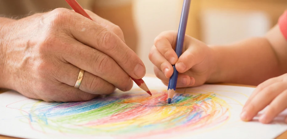Eine erwachsene Hand und eine Kinderhand, jeweils mit einem Buntstift, malen zusammen an einem Bild